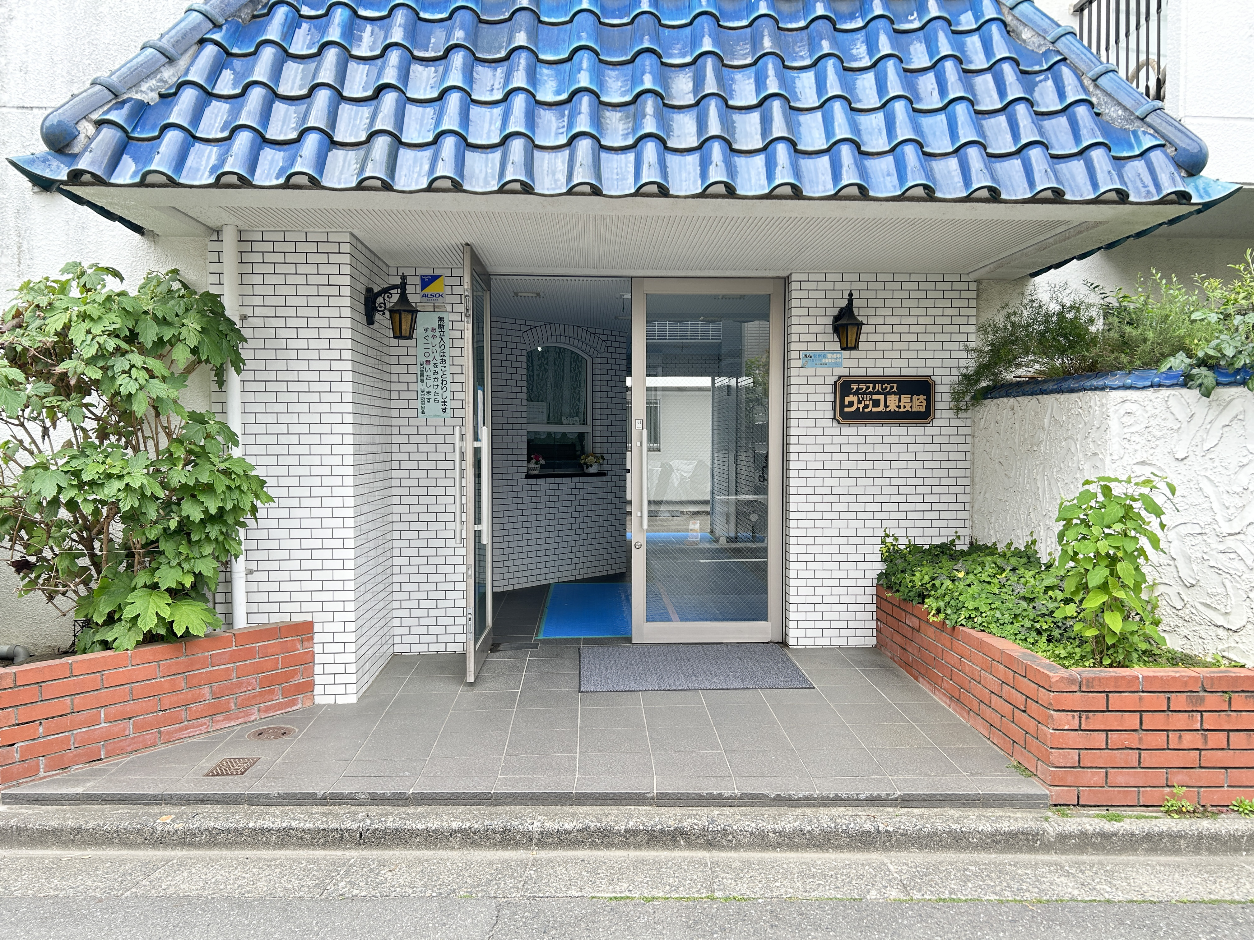 建物の玄関部分（ガラス扉、マット、植栽など）を主体に撮影した画像